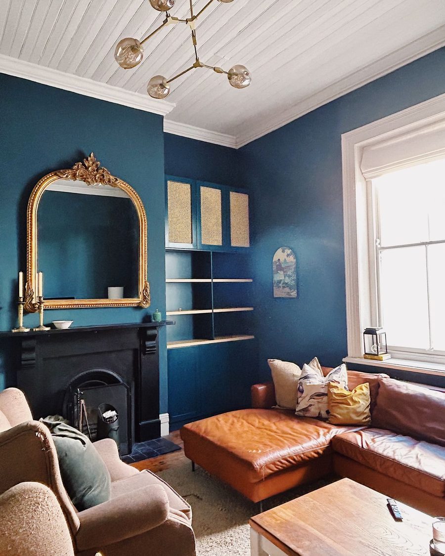 A heritage living room painted deep teal, with a gilded arched mirror above a black fireplace, built-in shelving and a tan leather sofa