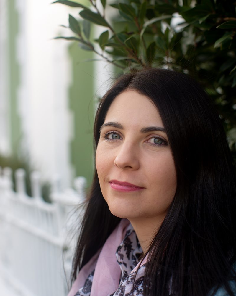 Portrait of Kirsten Burgess, smiling, beside a white fence and green foliage