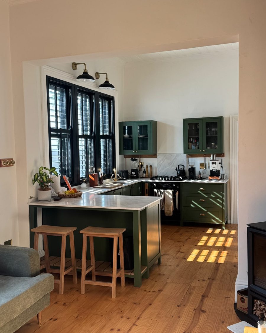 A heritage kitchen with sage green cabinetry, brass fittings and black-framed shutters, opening onto a pine-floored living room