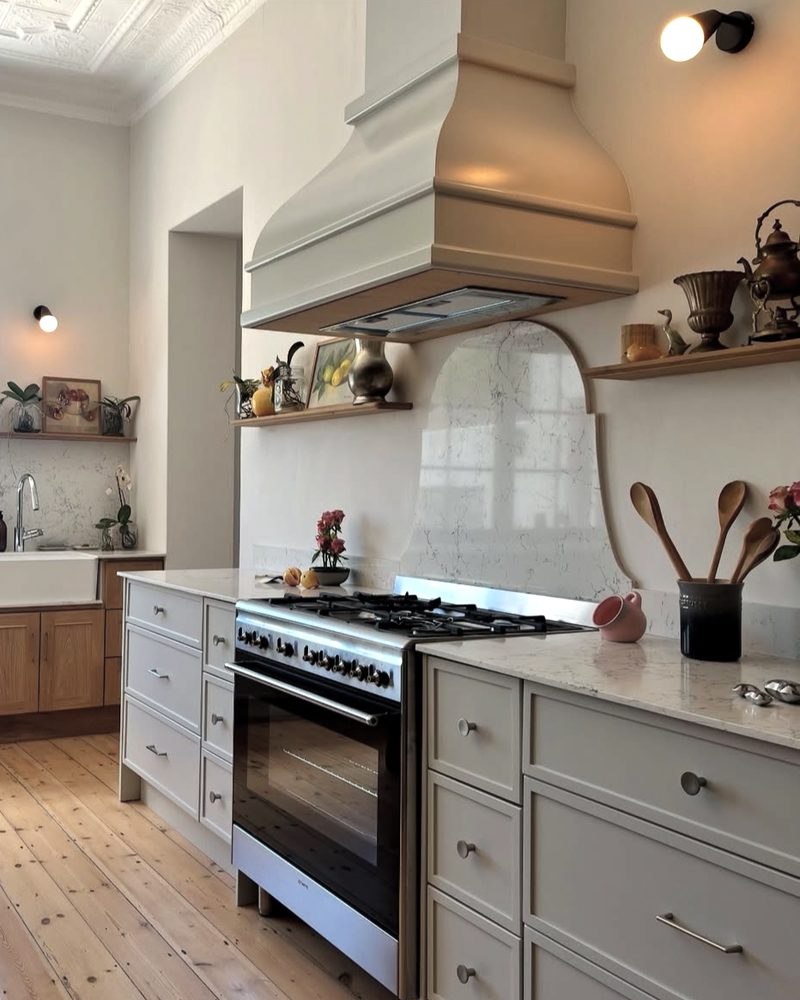 A heritage kitchen with a plaster range hood, an arch-shaped stone splashback, open timber shelves and pine floorboards